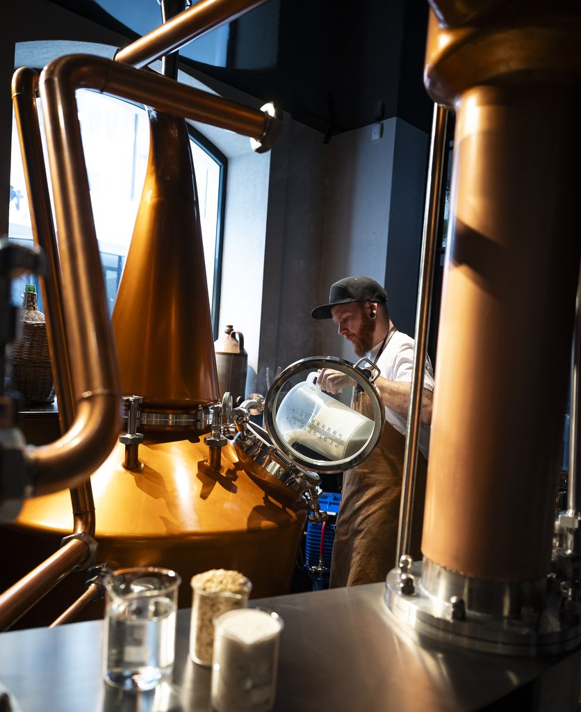 Distiller pouring milky liquid from a jug into a copper still sight glass