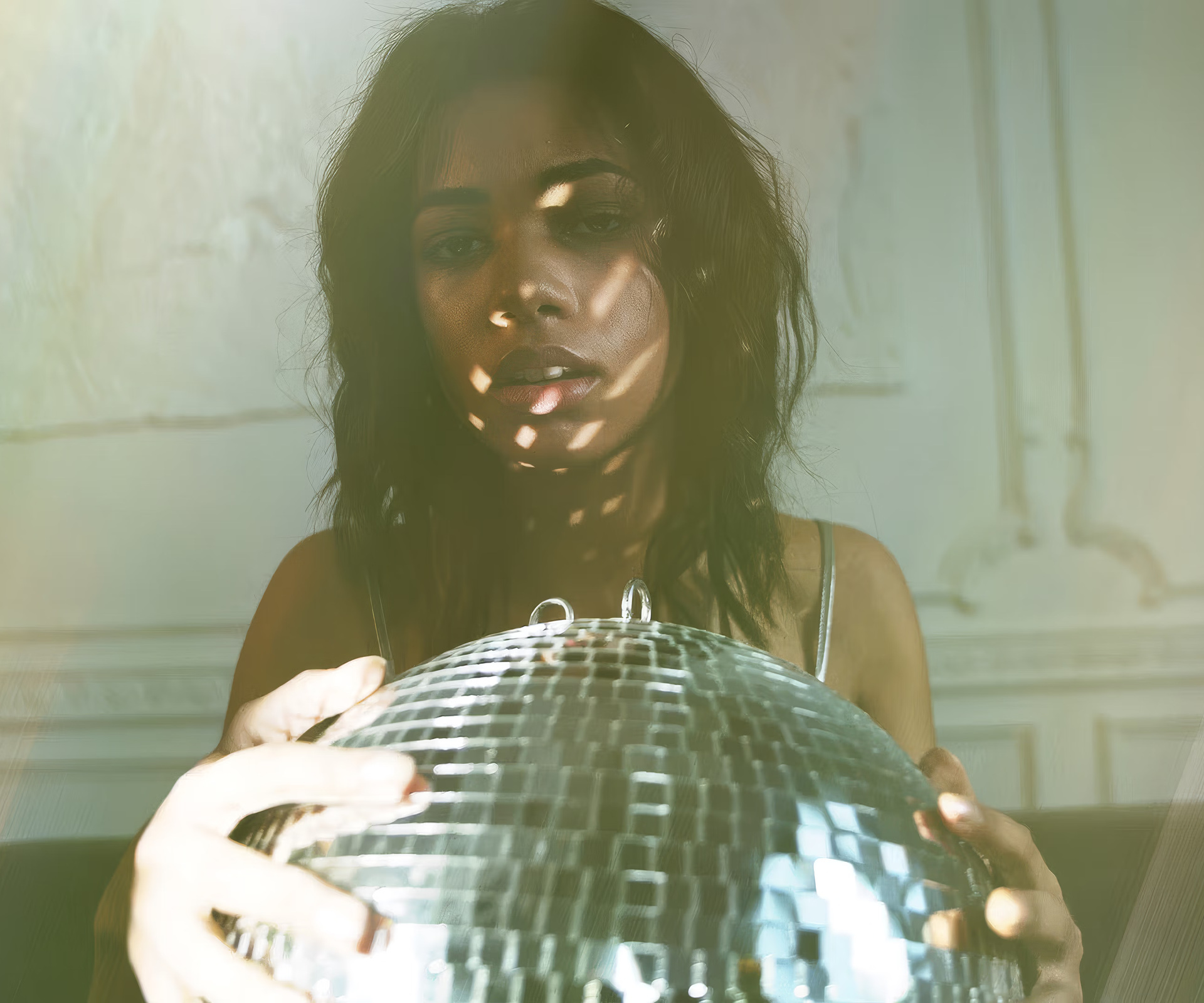 A woman with dark hair holding a mirrored disco ball, featuring dramatic striped shadows across her face and colorful vintage-style lens flares.